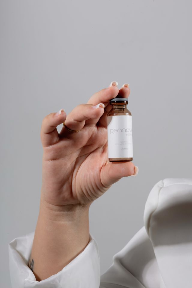 Hand holding a small amber glass vial labeled Rennova peptide serum, worn by person in white lab coat